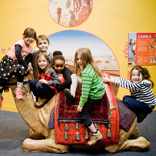 group of children sitting on a plastic camel with a yellow wall in the background