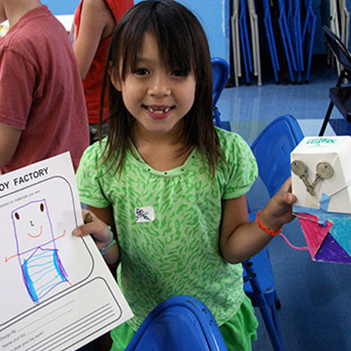 female child with black hair and green shirt holding a paper and science project