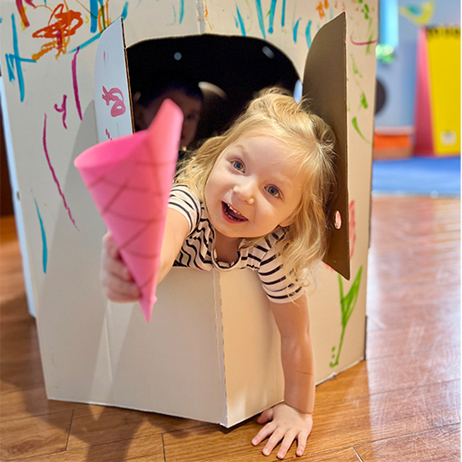 blonde female child leaning out of small structure on wooden floor holding pink paper in a conical shape