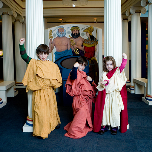 children wearing togas over clothes holding fists in air with columns in the background