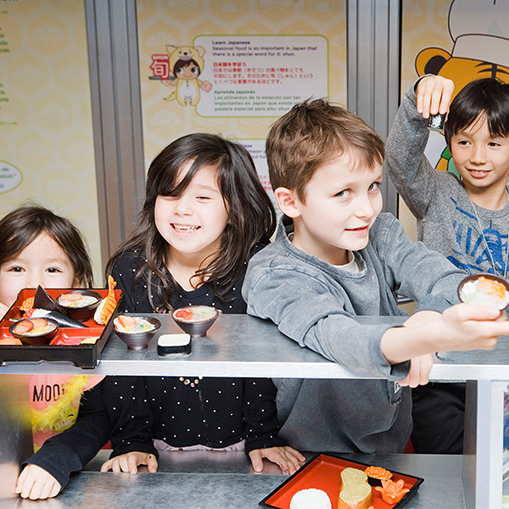 group of children behind a counter pretending to serve plastic sushi