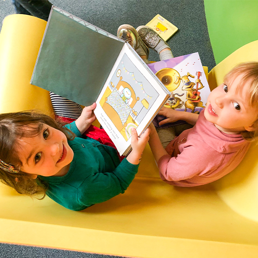 brunette female child and blonde child sitting in yellow chair together, each reading a book