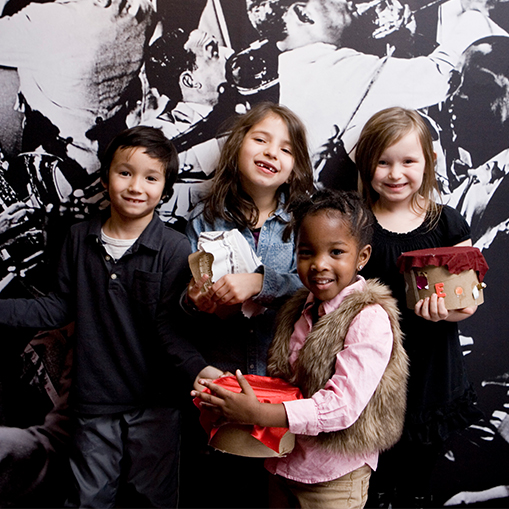group of children holding art projects standing in front of a black and white background featuring jazz musicians