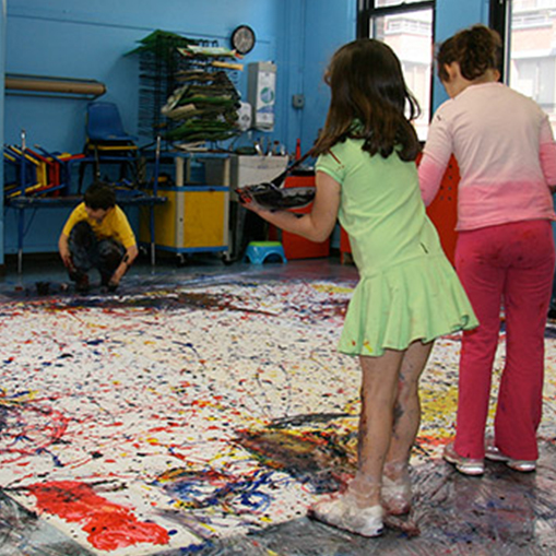 girl in a green dress with brown hair and girl in a pink top and red pants throwing paint onto the ground which is covered in paint