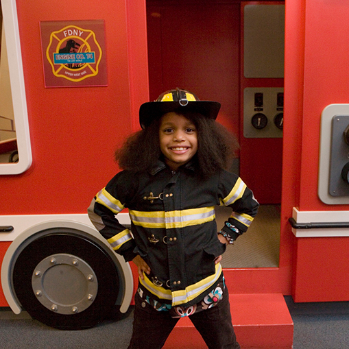 black girl dressed as a firefighter standing in front of a play firetruck