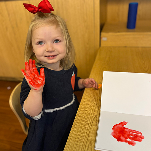 blonde female child with red ribbon in hair holding up hand with red paint on it. paper on desk in front of child with red handprint on it
