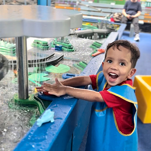 boy smiling in red shirt and blue smock holding hands out under cloud shaped sprinkler with water drizzling down on hands
