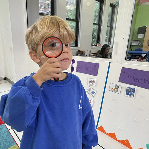 blonde male child in blue long sleeved shirt holding a magnifying glass to one eye