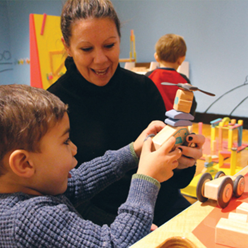 A boy builds a flying vehicle out of Tegu blocks with woman and other child in the background