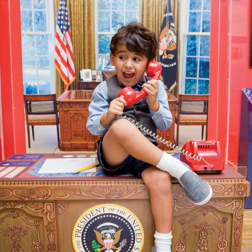 Boy sitting on miniature version of the oval office desk and speaking on a red phone smiling.