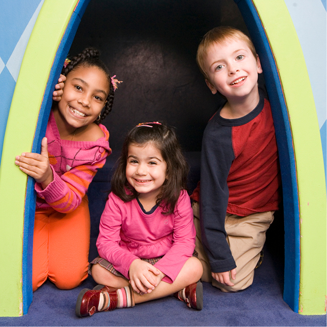 Three smiling kids peak out from a colorful doorway in the playworks exhbit.