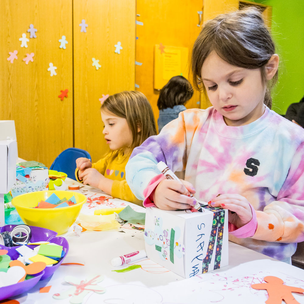 Small child in tie-dye shirt coloring a white cube with markers on a white table with other markers. Other small child next her with yellow top holding color paper.