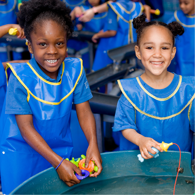 Two girls in blue smocks smile at the camera as they play in CMOM's H2O water exhibit.