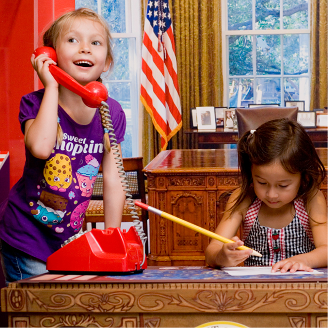 A girl pretends to speak on the phone while another girl pretends to write with an oversized pencil.