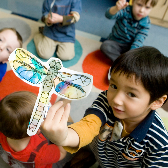 A boy looks up and holds up a dragonfly that he water colored.