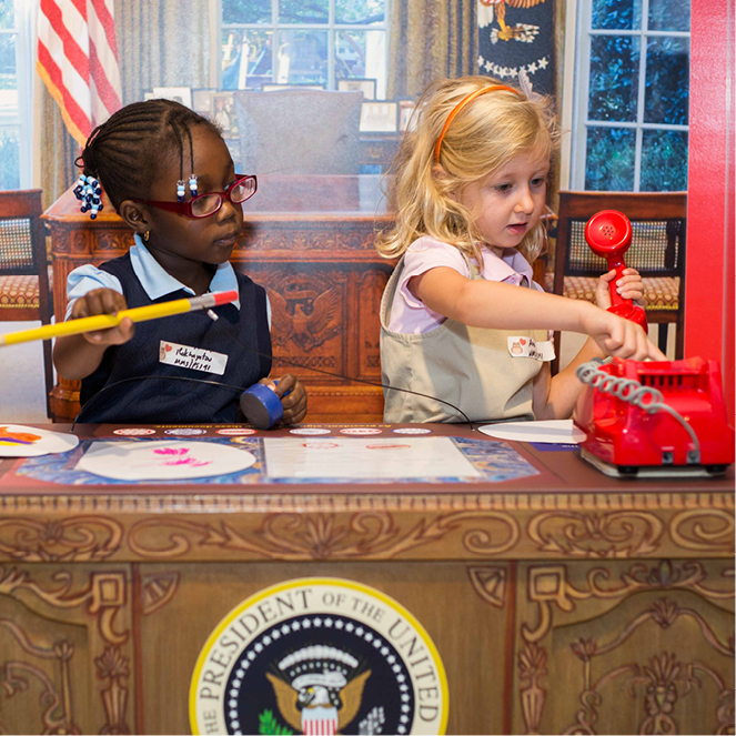 Two kids sit at a pretend oval office desk and pretend to write and speak on a red phone.
