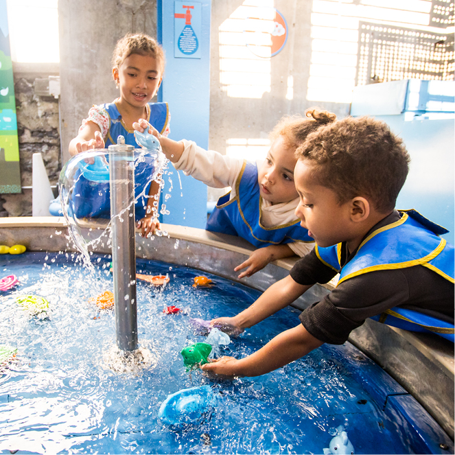 Three kids wearing blue smocks happily play on a sunny day in the outdoor Dynamic H2O exhibit.