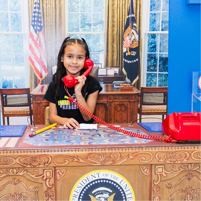 A girl sits at a pretend Oval Office desk and speaks on a red phone.
