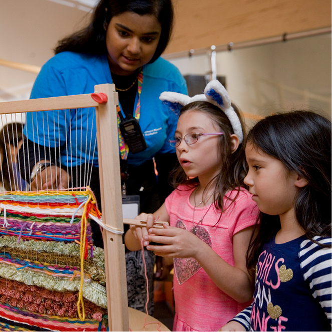 Two kids work on a colorful weaving with a CMOM educator