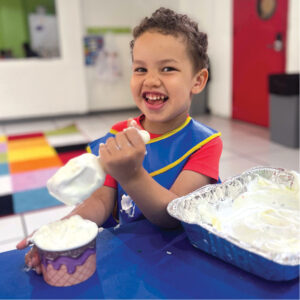 A boy wearing a blue smock smiles widely and scoops shaving cram into an ice cream cup