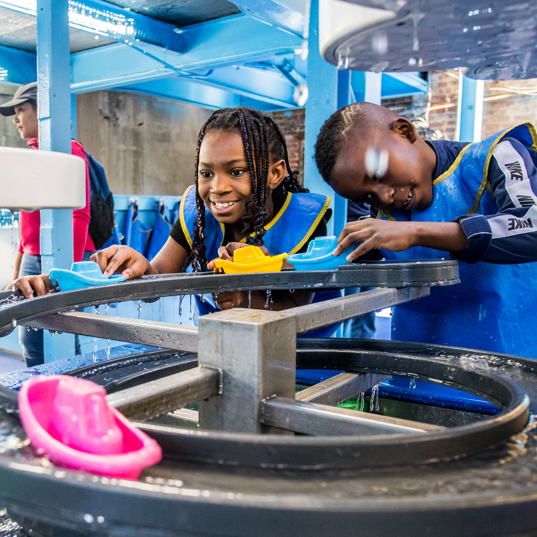 Multiple children in blue and yellow water smocks with plastic boats putting them into a water exhibit.