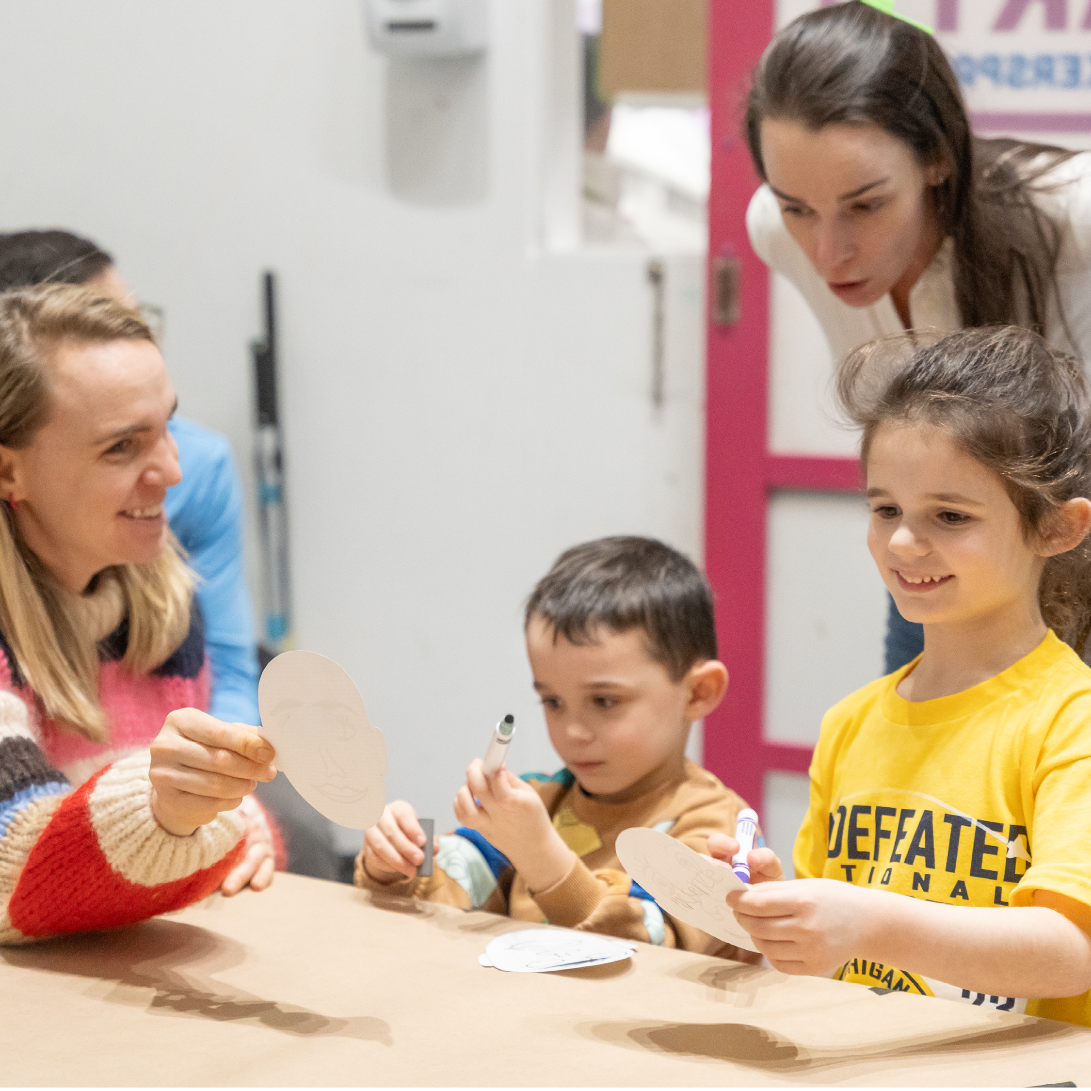 A caregiver holds up their self portrait as two children and another caregiver smile and look on.