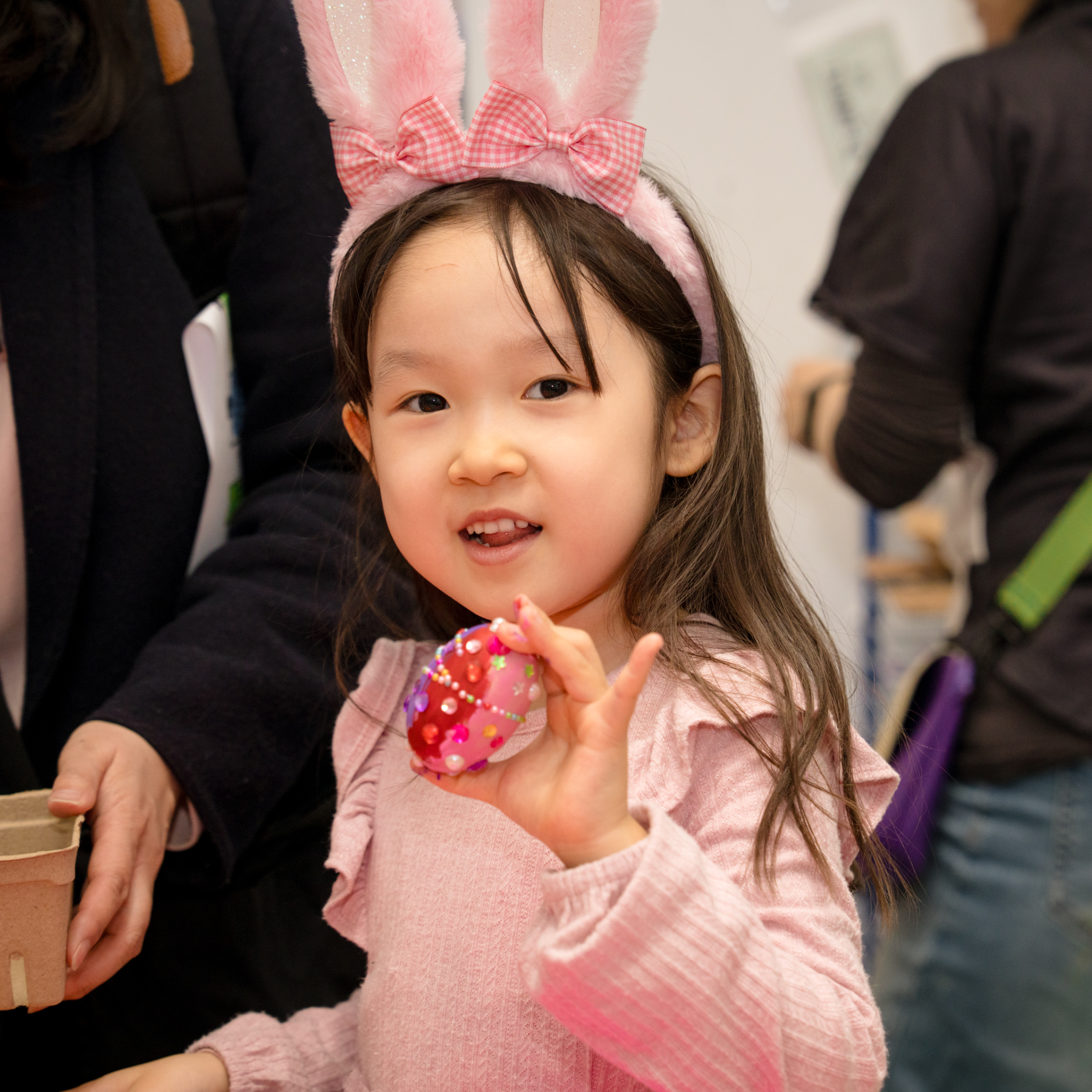 A child wearing pink bunny ears happily holds up a decorated Easter egg.