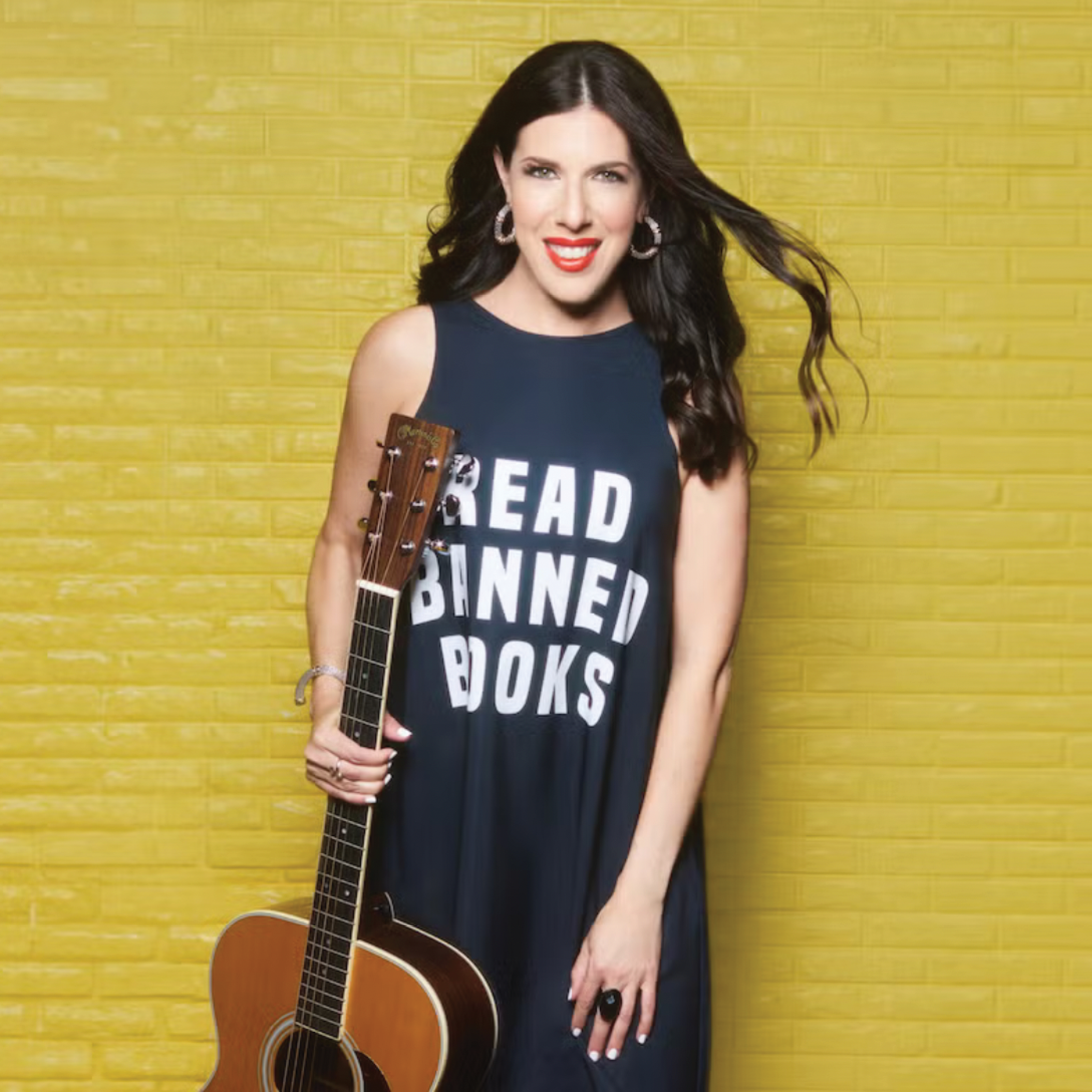 Singer song writer, Joanie Leeds, stands against a yellow background smiling and holding a guitar.