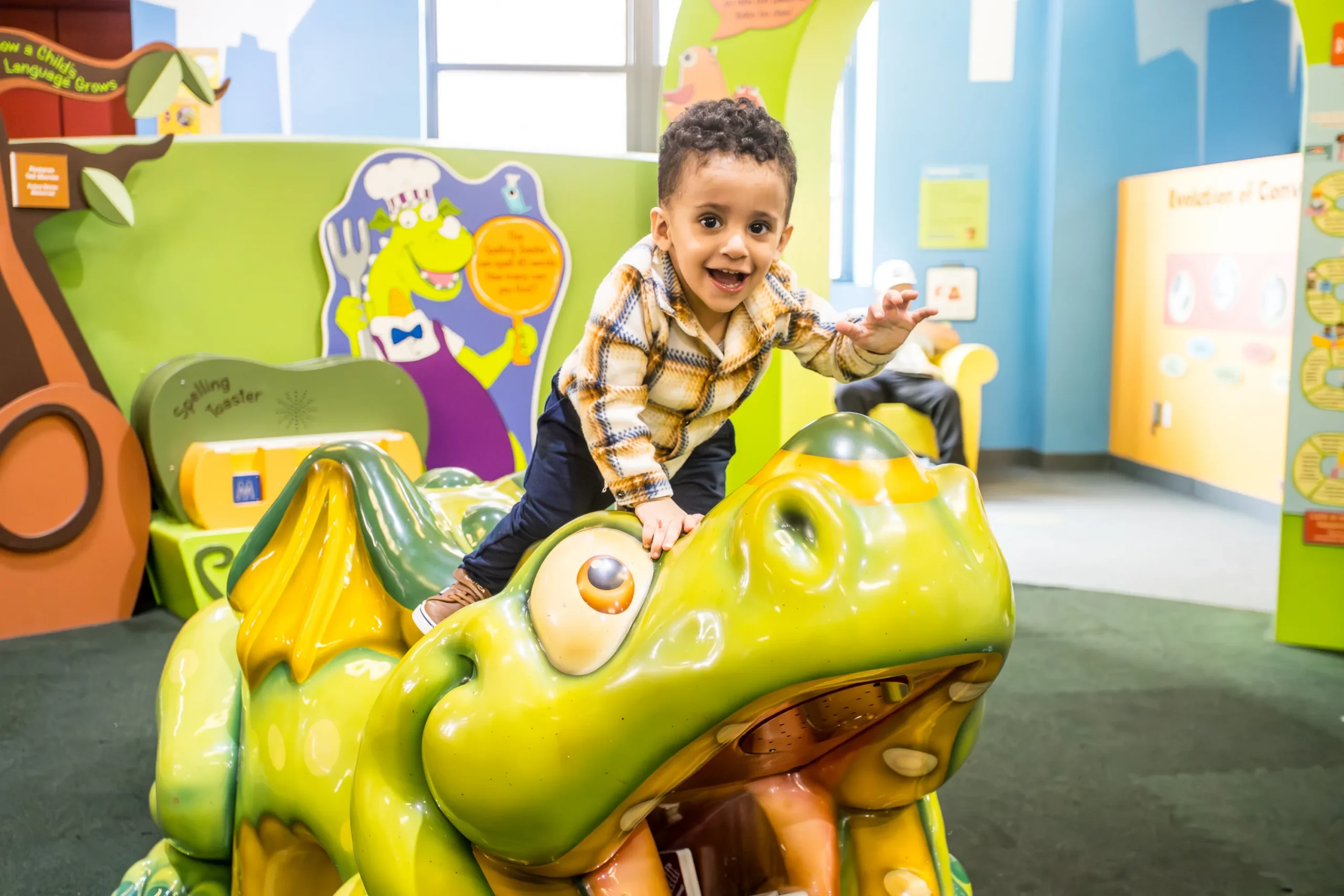 A smiling young child climbs a figure of a dragon named Alphie, the unofficial mascot of Children's Museum of Manhattan