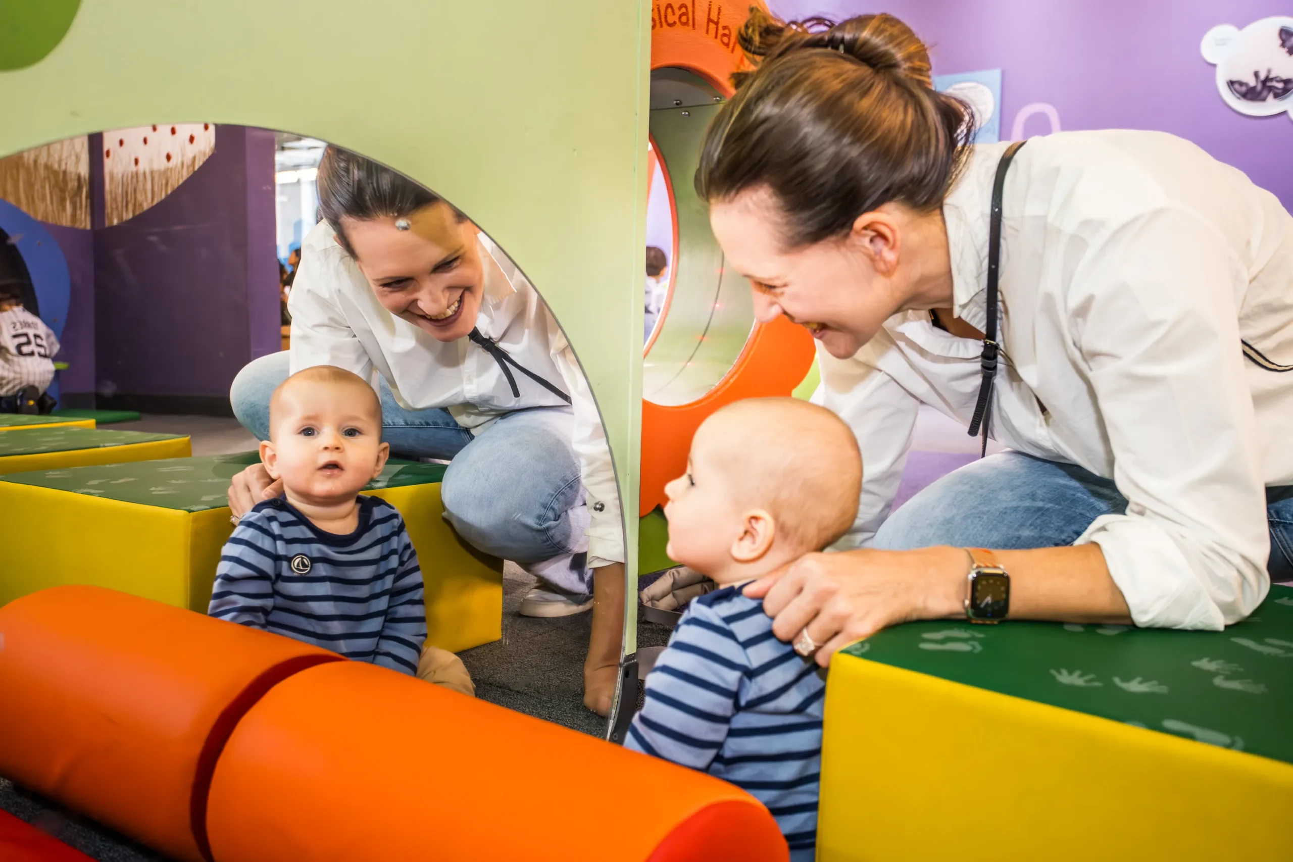 An adult woman smiles at a baby, who sits in front of a mirror. The reflection of the baby and woman are also featured.