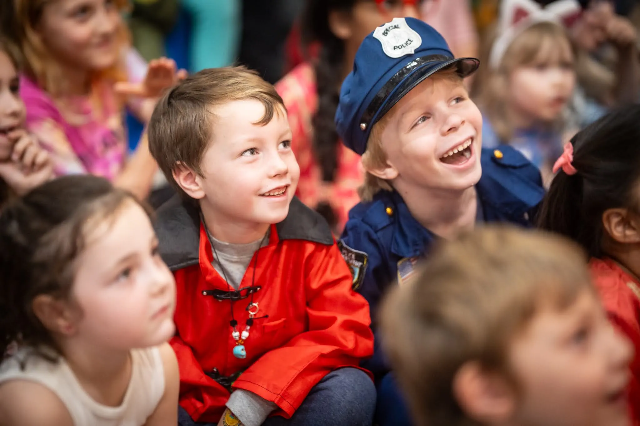 One child in a red jacket and another who appears to be dressed in a police officer's uniform costume, both smiling at something happening off-screen