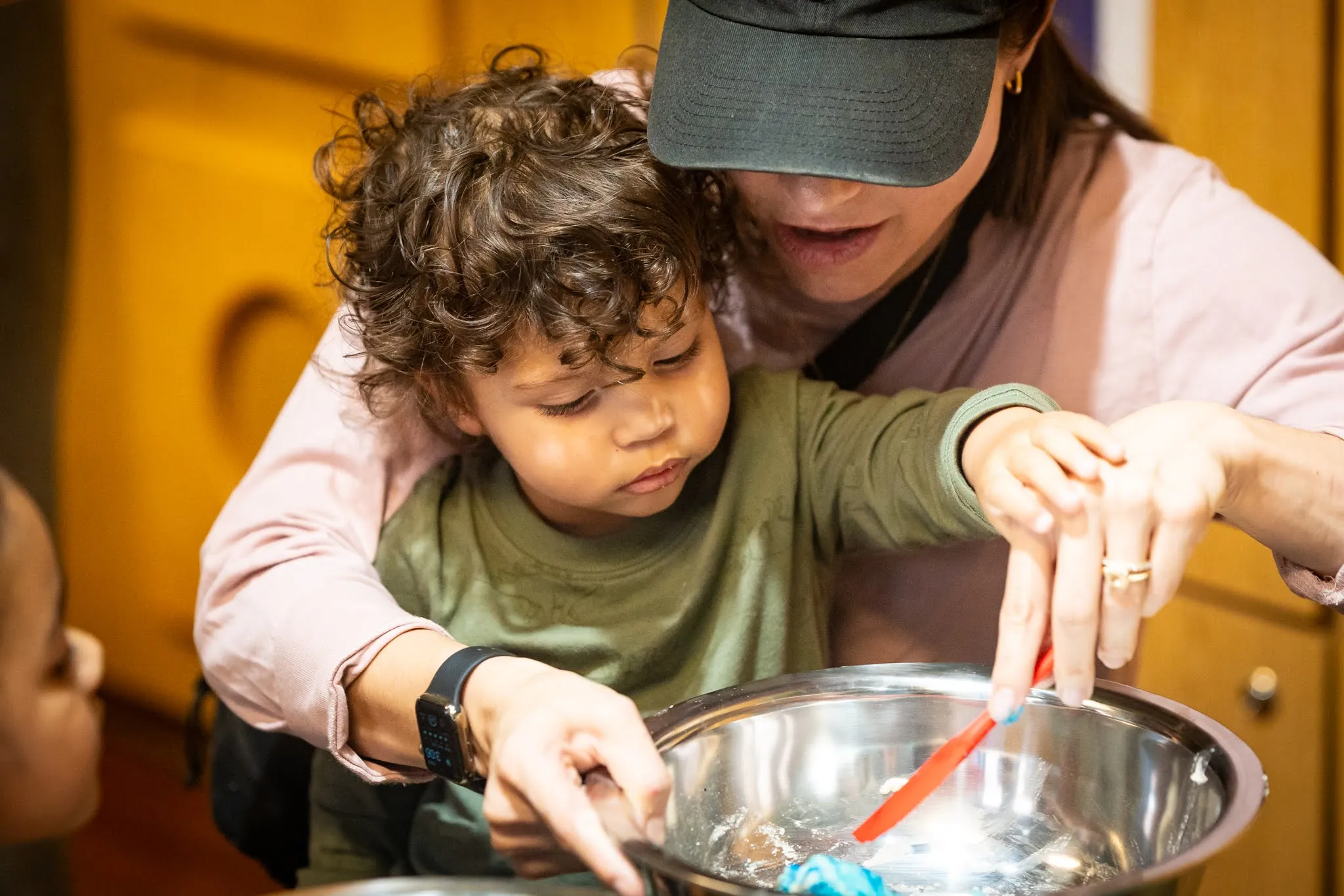 A young child gets assistance from an adult, who is wearing a baseball cap pulled down over their face, as the two stir something in a bowl