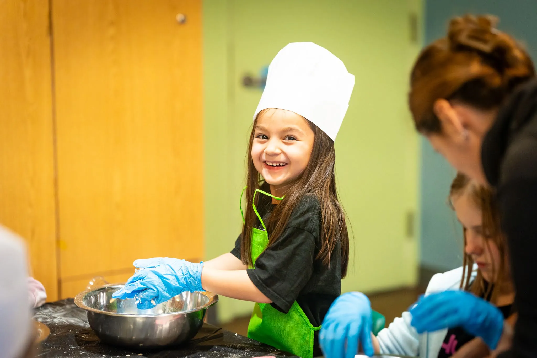 A smiling child in rubber gloves, an apron, and chef's hat, mixing together ingredients in a bowl