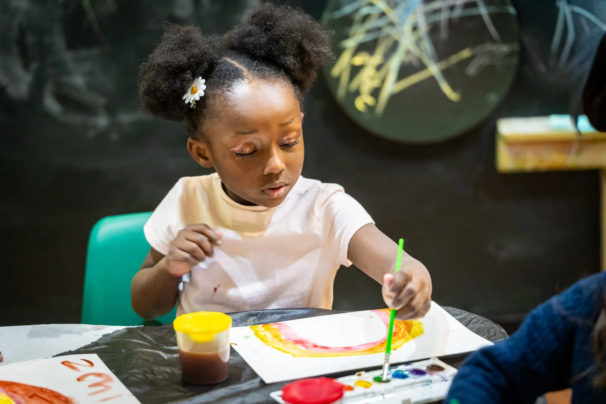 A small child dips her paint brush into green water color paint while she appears to be painting a rainbow on a piece of paper that is in front of her