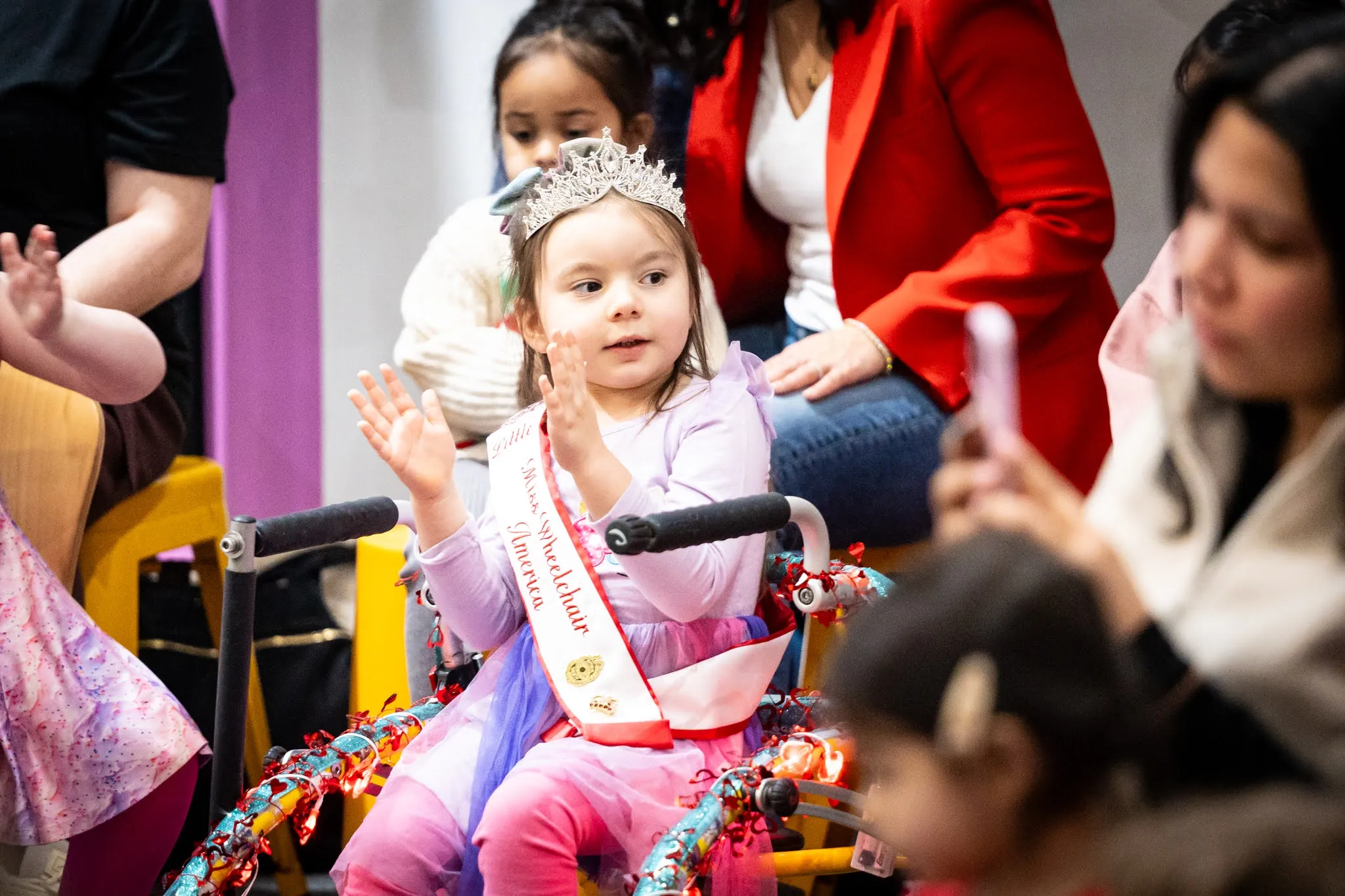 A child appears to be sitting in a child's wheelchair. The child wears a plastic crown and a sash that appears to say, "Miss Wheelchair America". The child is mid-clap.