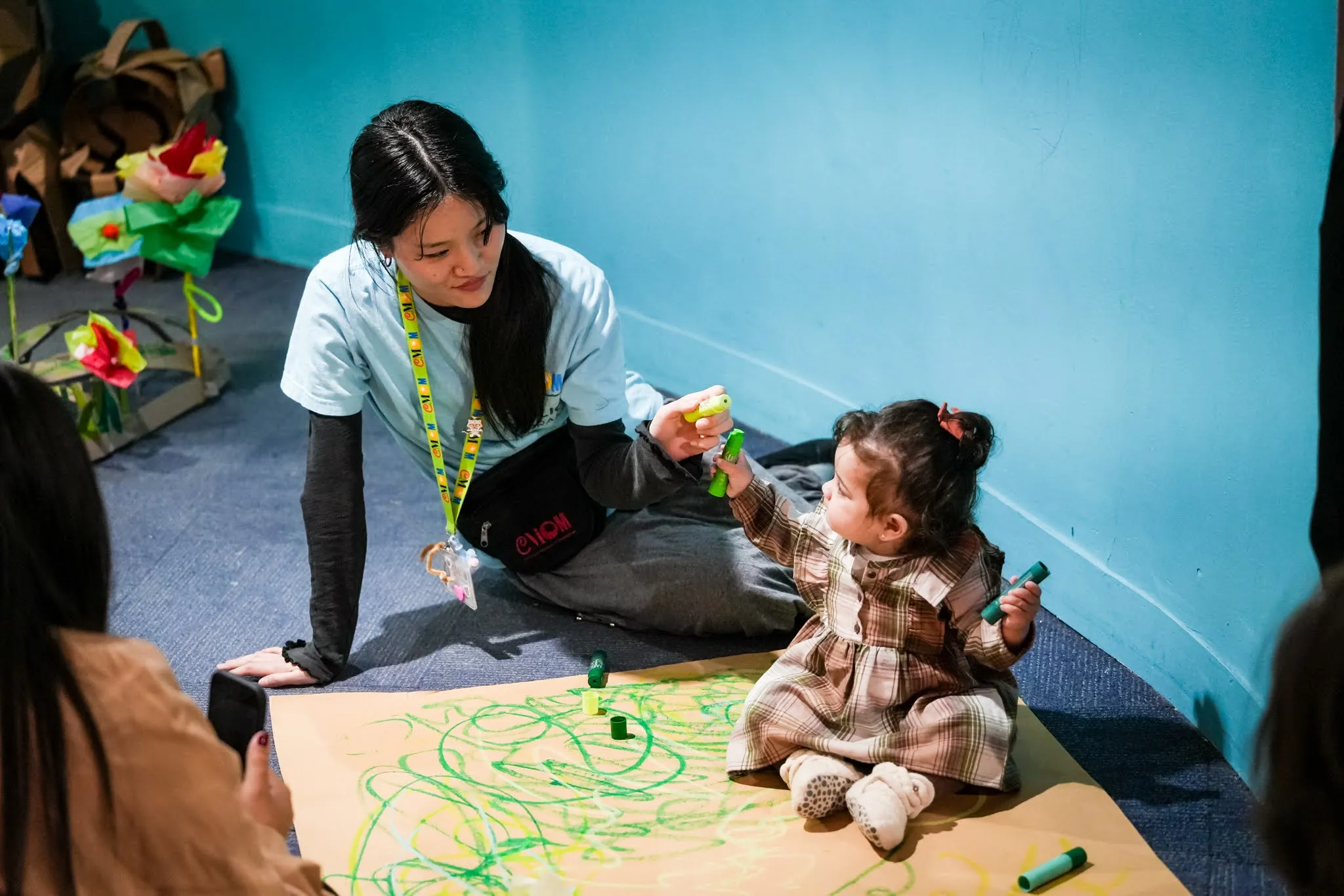 A young adult wears a nametag and uniform, signifying her as being a staff member at Children's Museum of Manhattan. She holds a marker up to an infant, who holds two markers. THe two appear to be coloring on a large paper in front of them, and the infant sits on the paper.