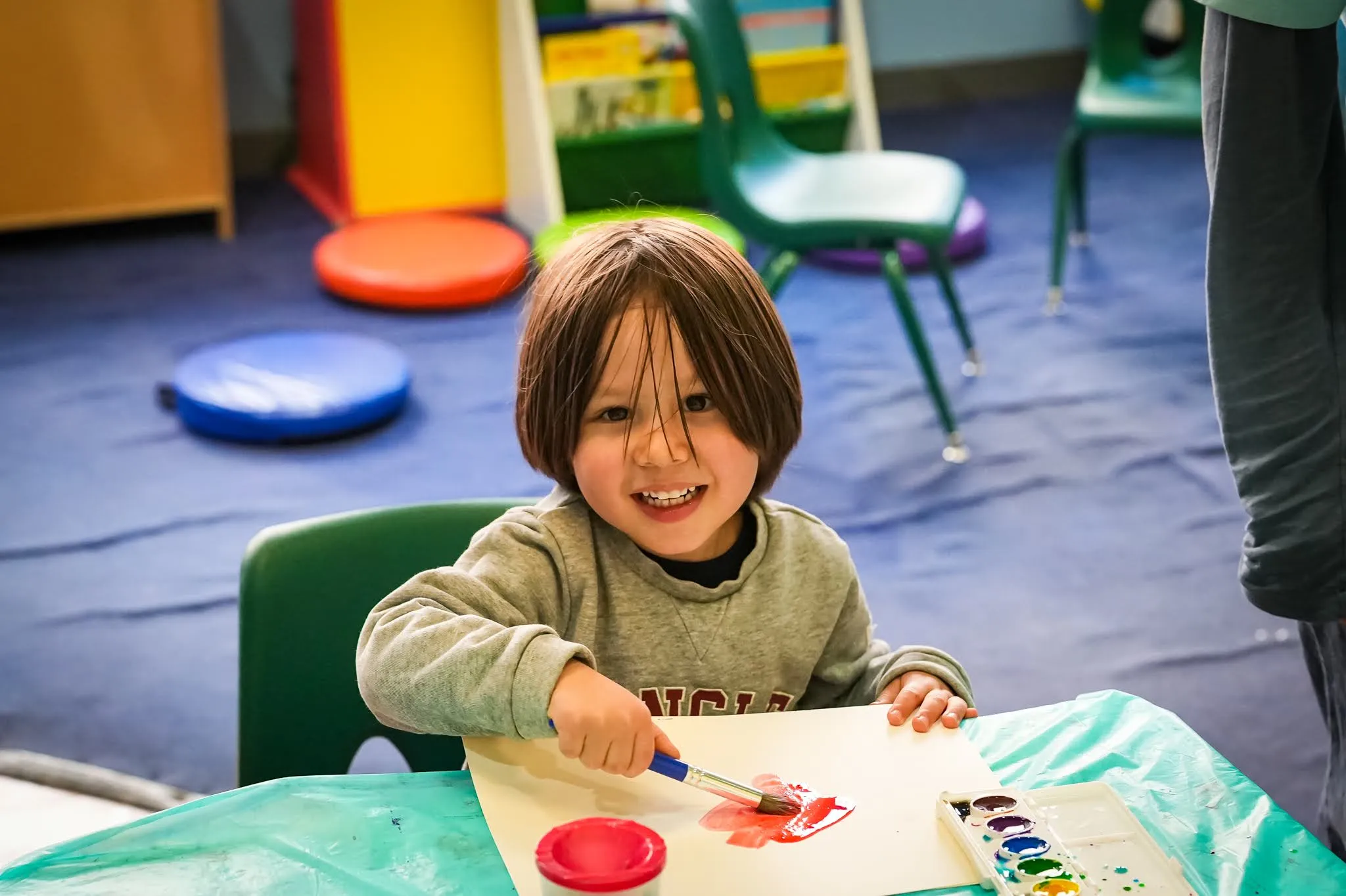A young child smiles at the camera while painting red paint on a paper in front of him on a table.