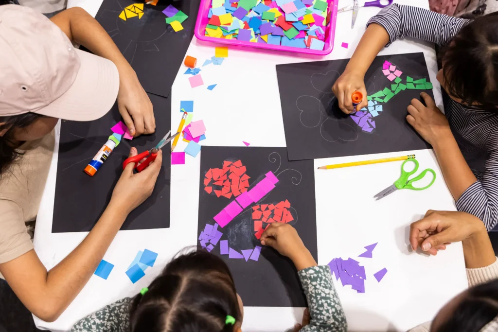 What appears to be children are shown crafting from above. They are assembling small pieces of cut-up paper, similar to mosaics.