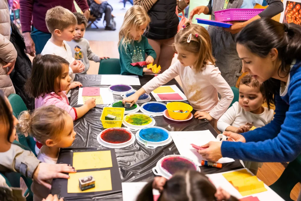 A large craft table with paints, and a group of children is sitting at it
