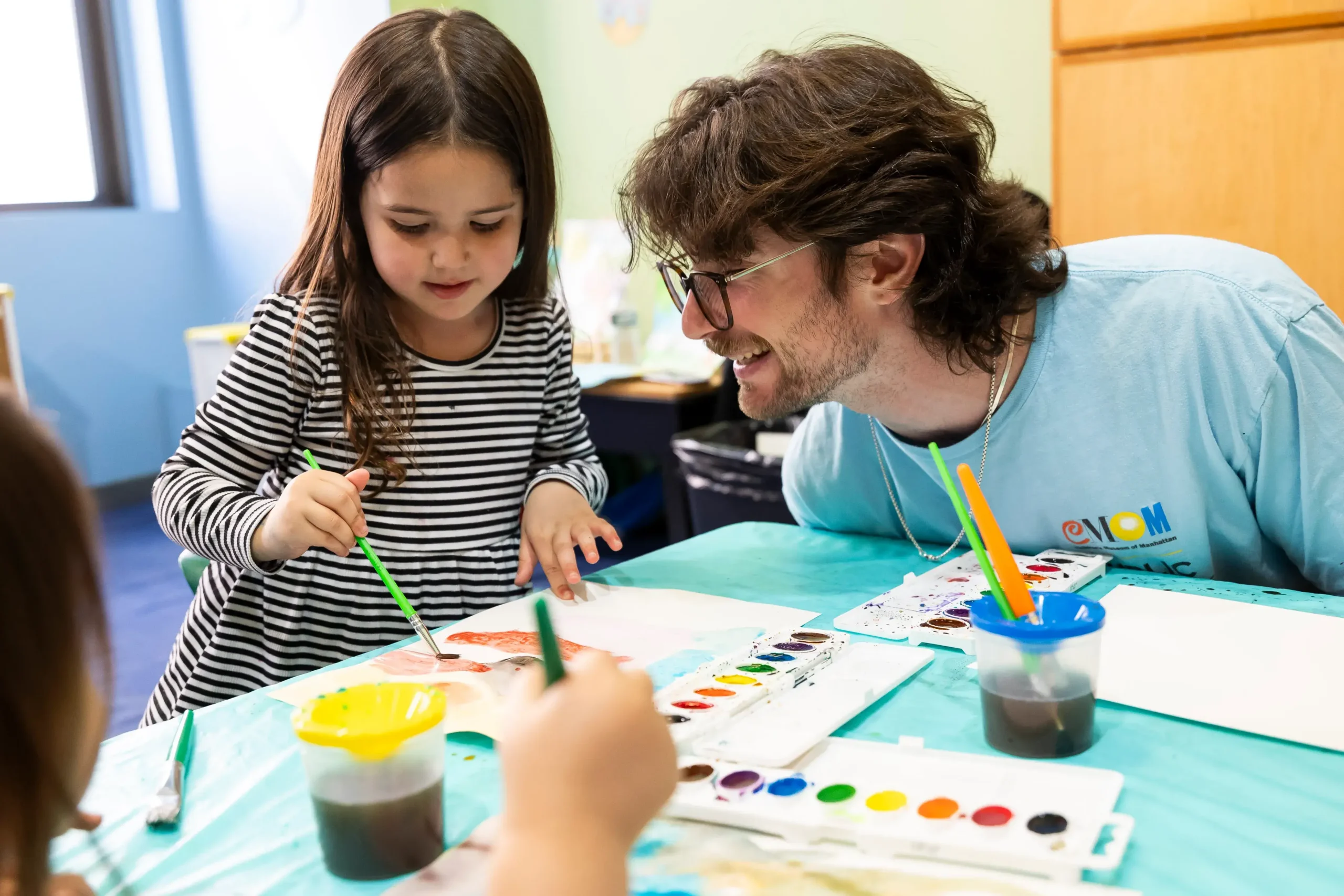 A young child paints on a paper in front of her, while a young adult sits at the table and smiles at her craft. The young adult wears a "Children's Museum of Manhattan" staff shirt.