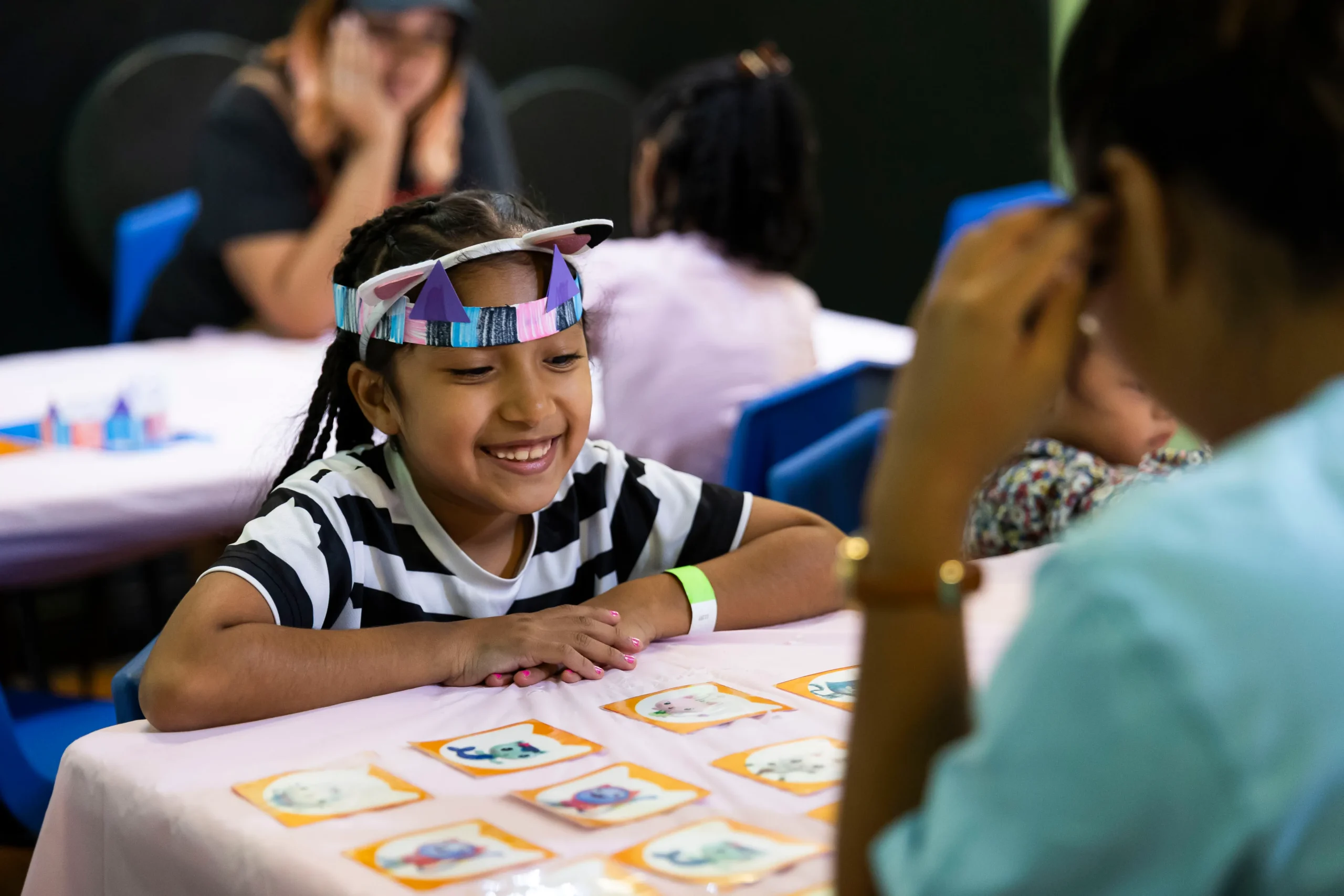 A young child in two different play costume headbands smiles while playing a card game with a person who seems to be an adult, who is in the foreground to the side and out-of-focus.