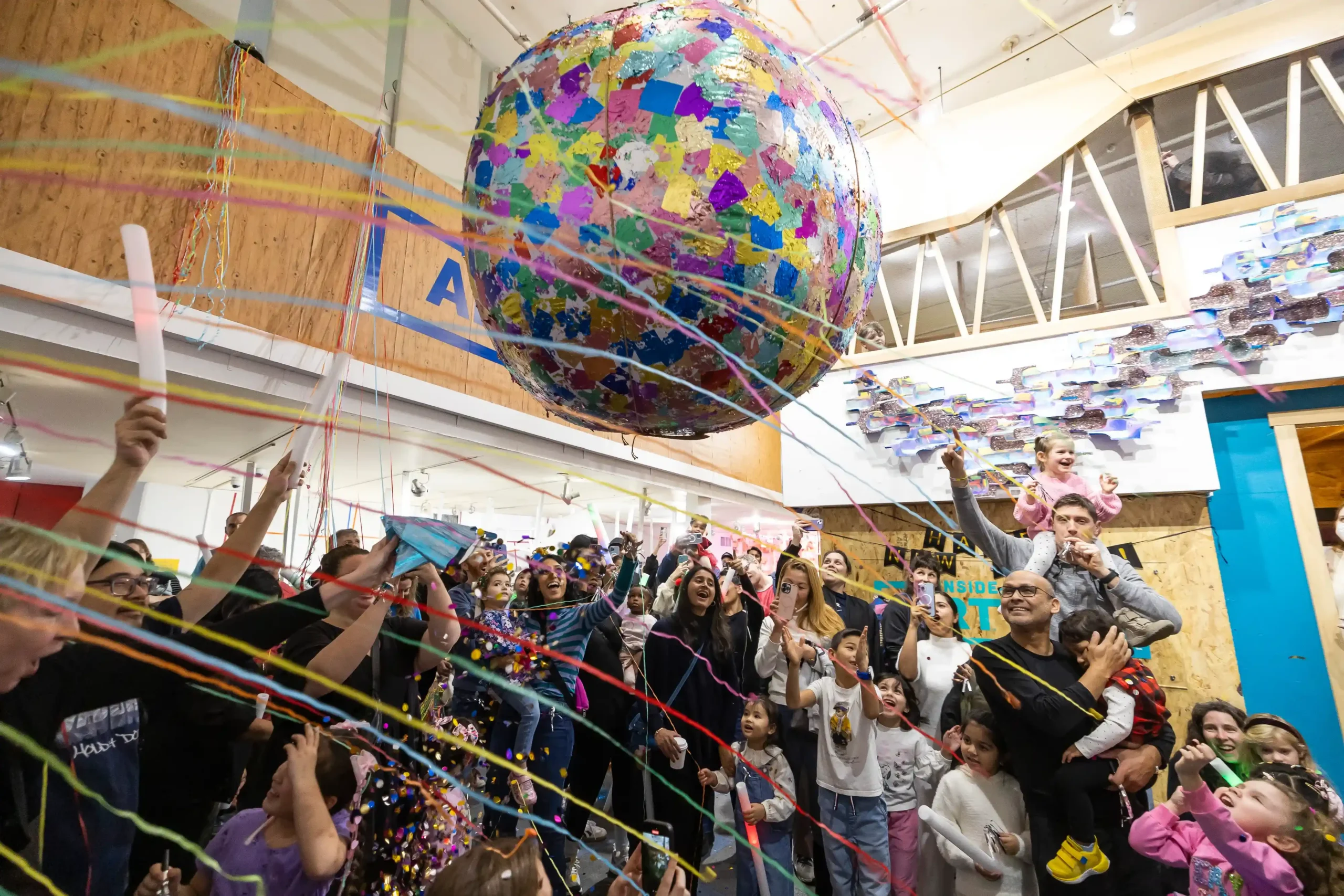 A colorful globe or orb is suspended from the ceiling, and colorful streamers go past. A crowd of adults and children cheer.