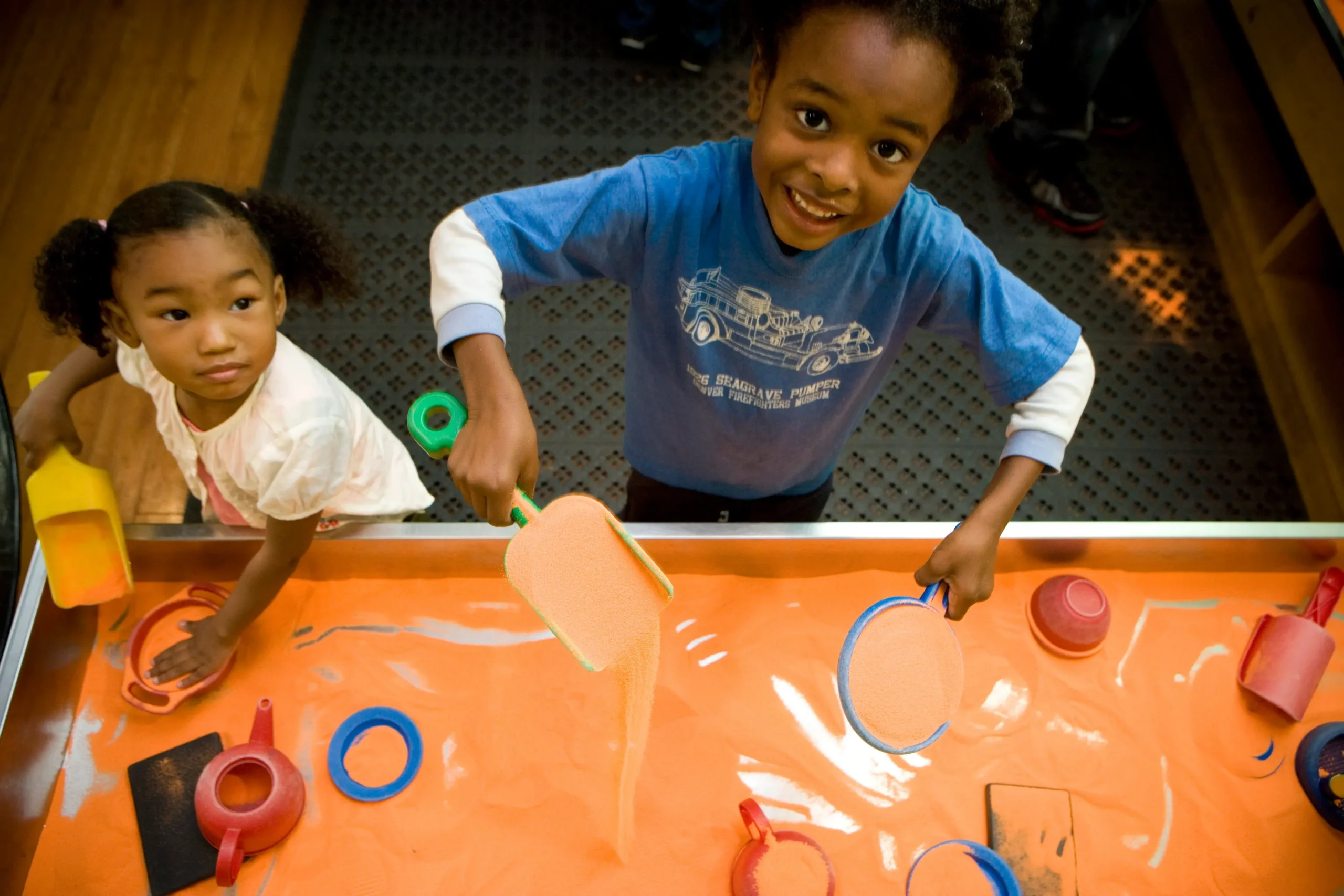 Two children play with shovels in a sandbox filled with bright orange sand. One of the children is looking at the camera and smiling.