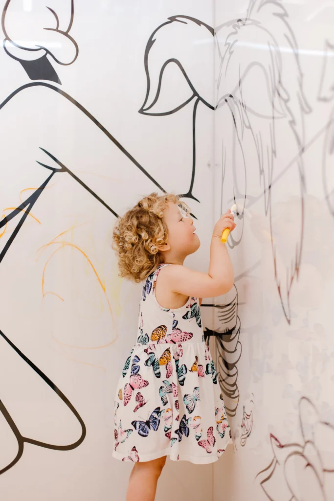 A child holds a marker and appears to be coloring in a black-and-white mural, which has more scribbles on the wall.