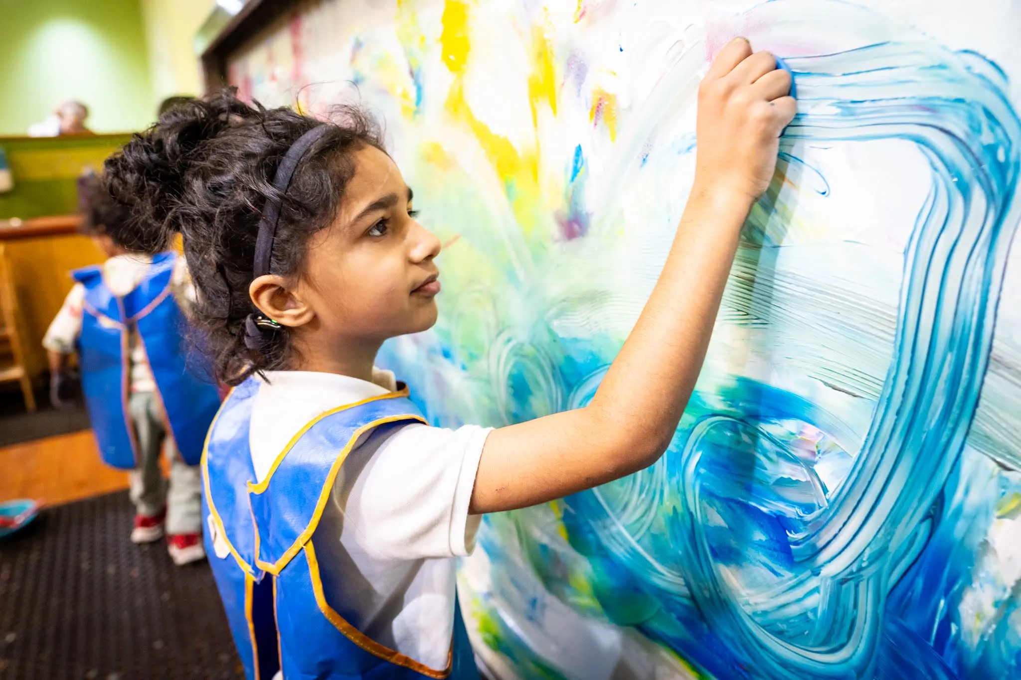 A child wears a smock and paints blue paint on to a large painting in front of her.