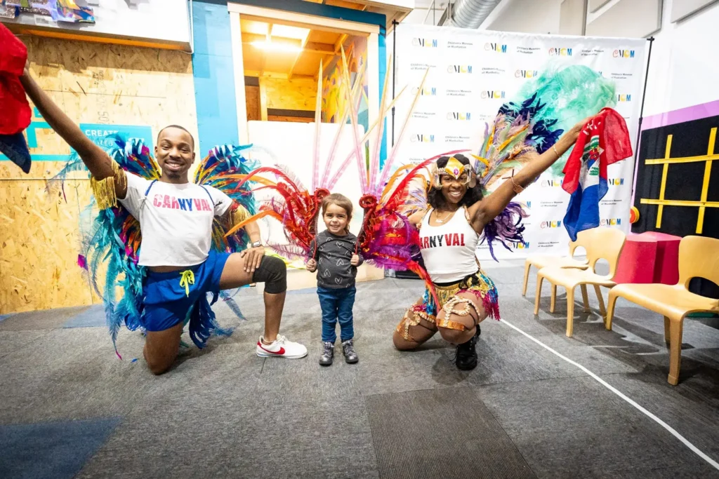 Two dancers wear shirts that say "CarNYval" (with NY in different color) and wear feathered costume accessories. They flank a small, smiling child who is holding more feathers.