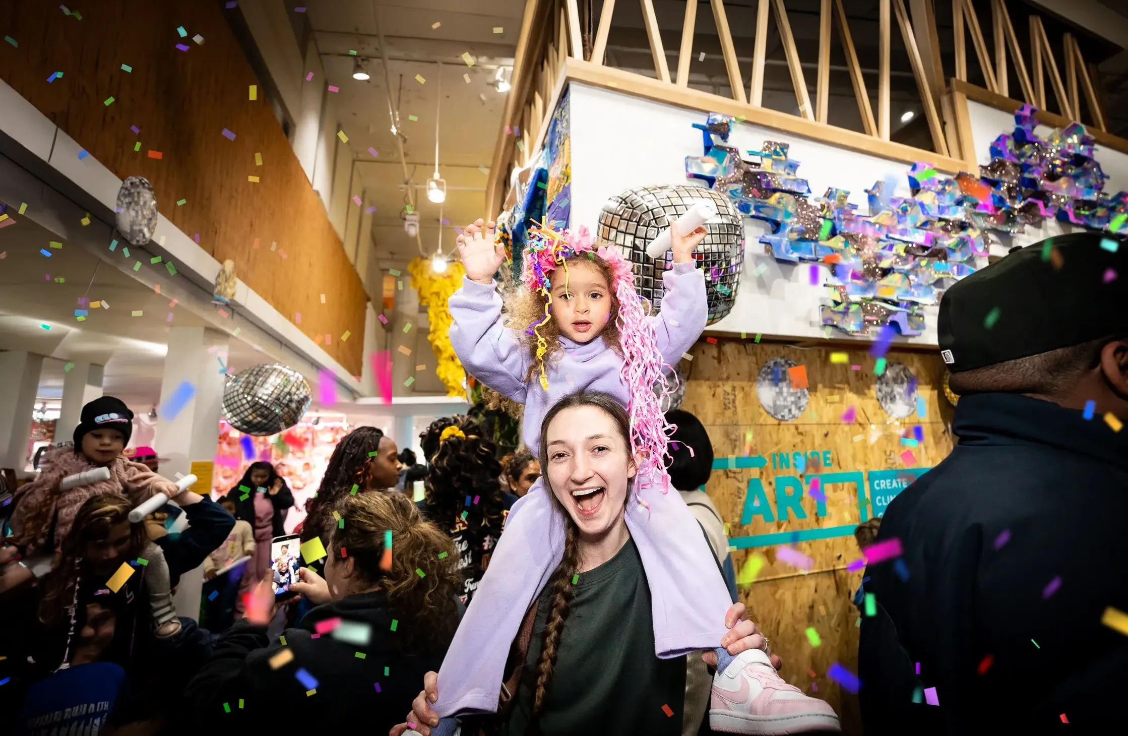 A young child with pink confetti in her hair sits on the shoulders of an adult. Both smile at the camera. In the background there is confetti and a crowd of people.