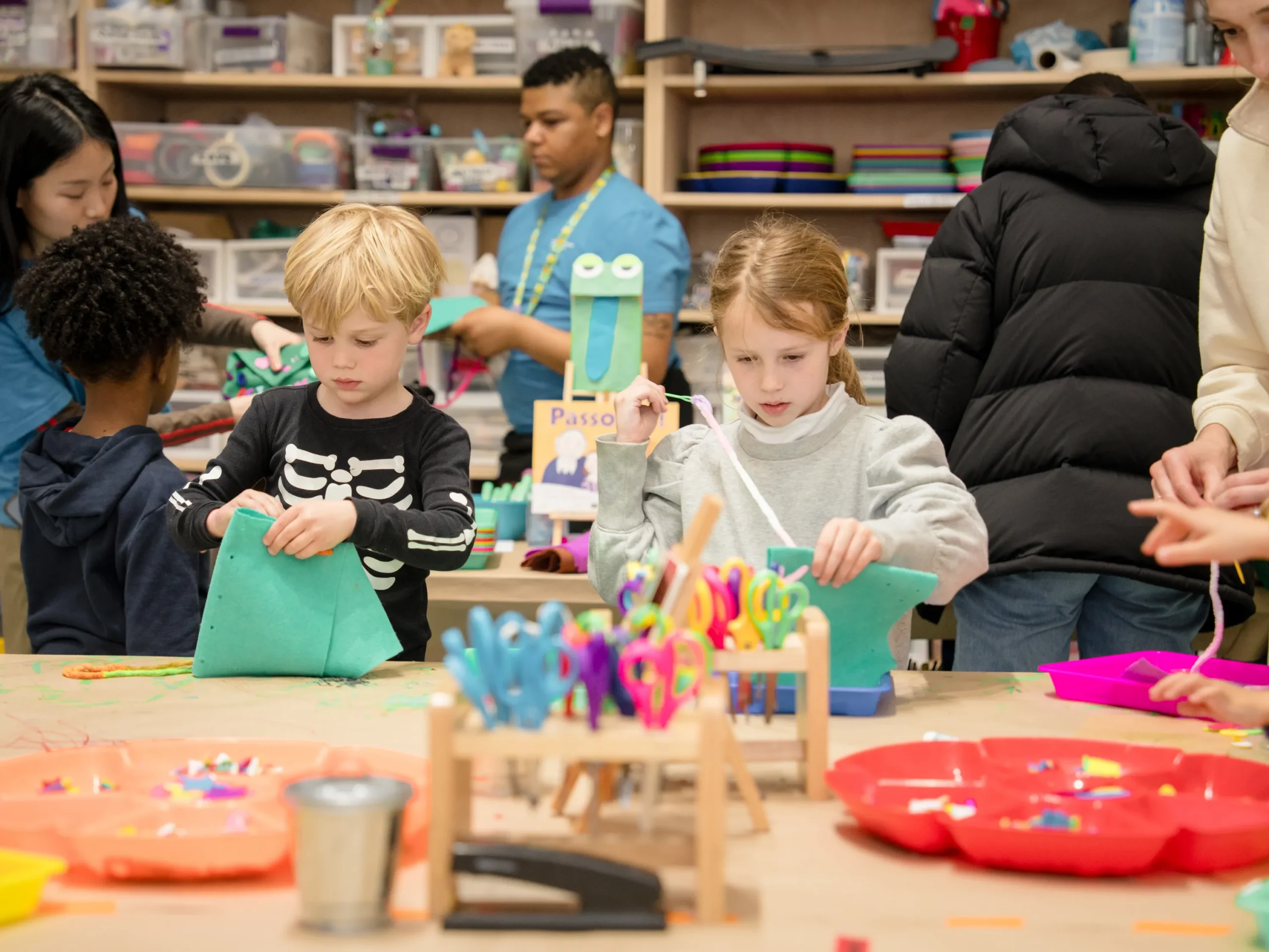 Two children work on paper crafts at a craft table with craft supplies on it, including many pairs of colorful scissors. In the background there is at least one additional child and perhaps three adults.