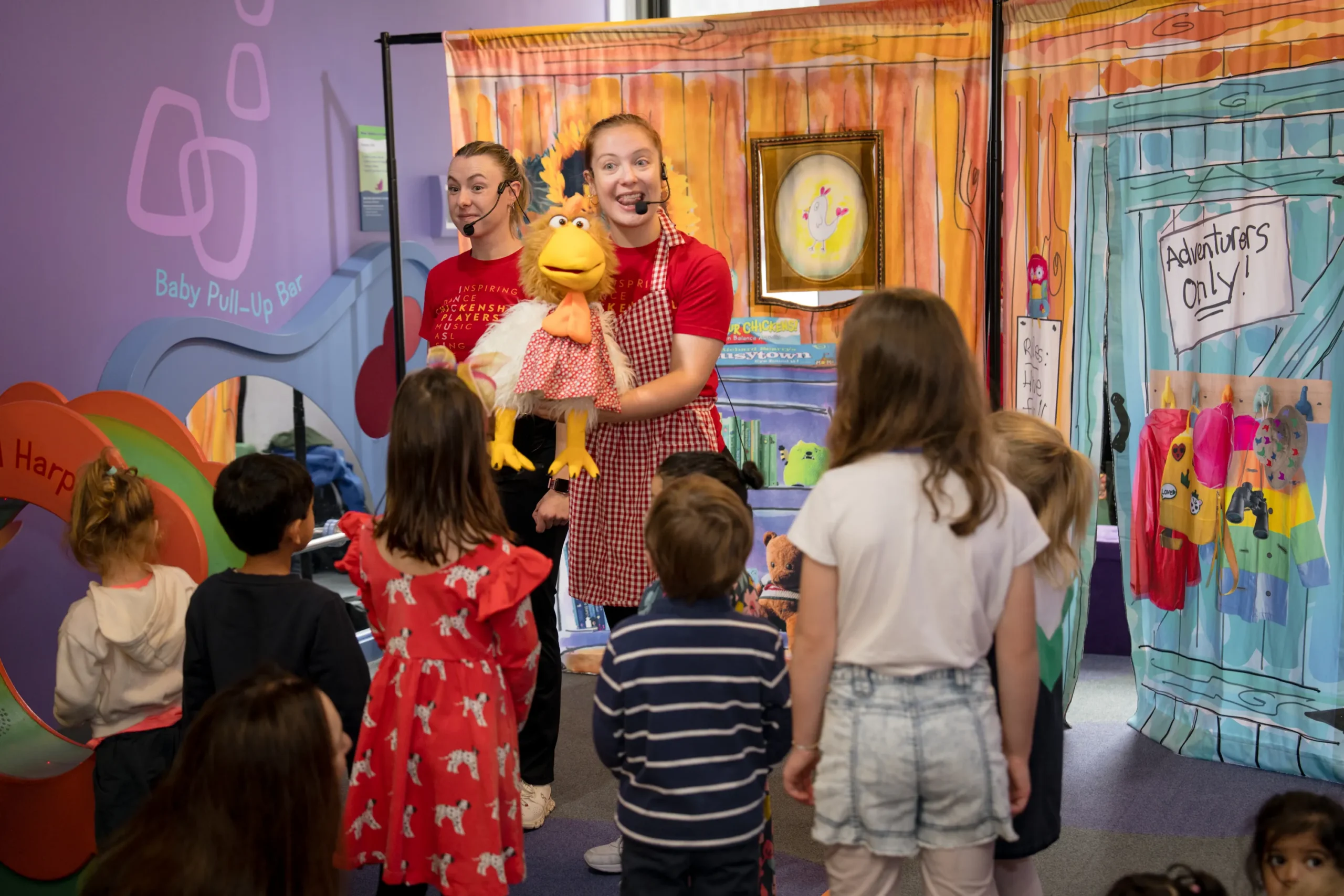 Two performers wear headset microphones and perform for a small group of children, who have their back to the camera. One o fhte performers holds a puppet of what appears to be a chicken. They are in front of a screen that has an illustrated door on it, with a sign that says "Adventurers Only!", underlined twice.