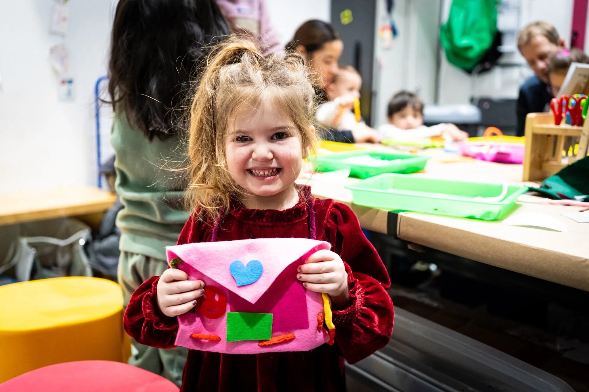 A smiling child holds up what appears to be a crafted felt purse or pouch that she has made. She is smiling at the camera.